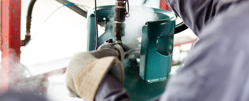 A person wearing protective gloves works with the valve of a teal propane tank while gas releases around the valve