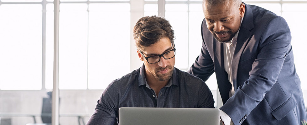 One businessman sits and looks at a laptop while a manager stands next to him, displaying a coaching manager posture.