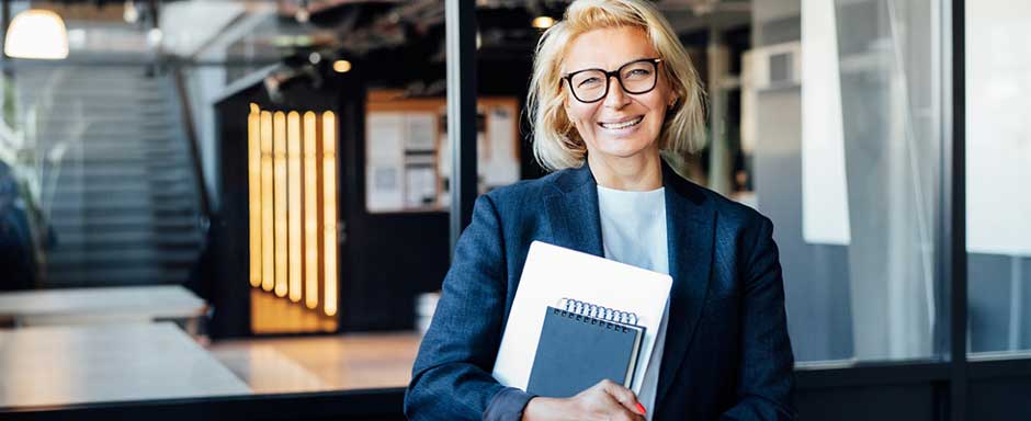 A professional woman in glasses smiles and hold a notebook in an office setting