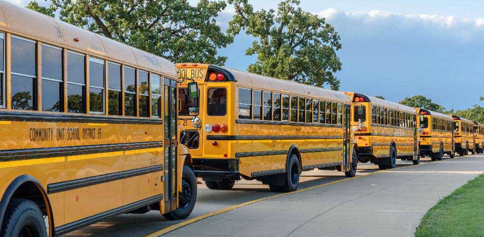 Six yellow school buses lined up down a paved road with cloudy skies and green trees in the background