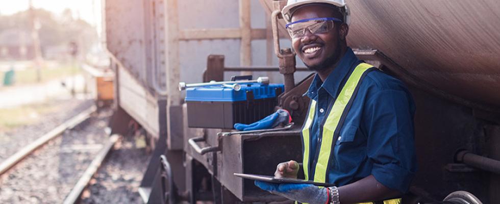 A man in a safety vest poses with his back against a train and a clipboard in his hands.